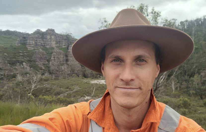 RPS environmental scientist Tam Durney wears orange high-vis PPE and a brown Akubra hat - behind him is a rocky mountain cliff. 