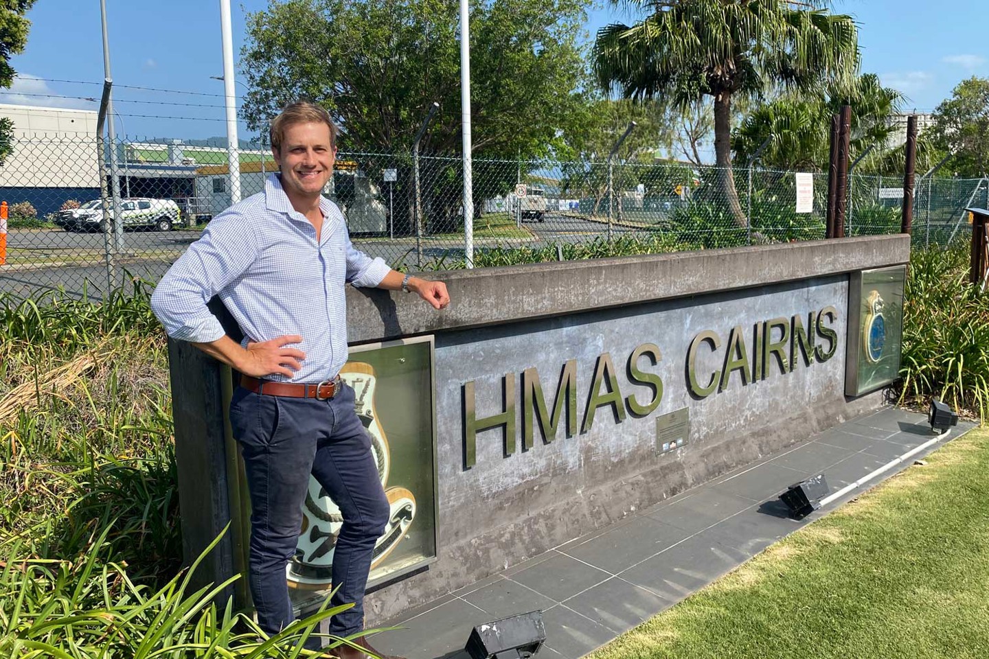 RPS Project Manager, Lee Leitner stands in front of a HMAS Cairns sign surrounded by grass and shrub. He wears a blue shirt and jeans and has his right arm in a pose. 