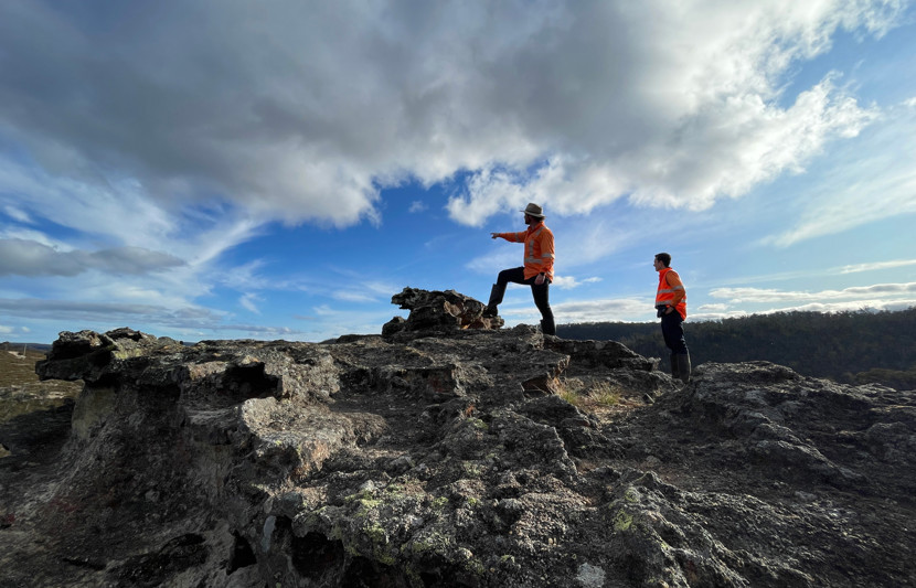 Two RPS environmental scientists dressed in orange high-vis stand on rocky terrain with one pointing to a distance as the clouds sweep overhead. 