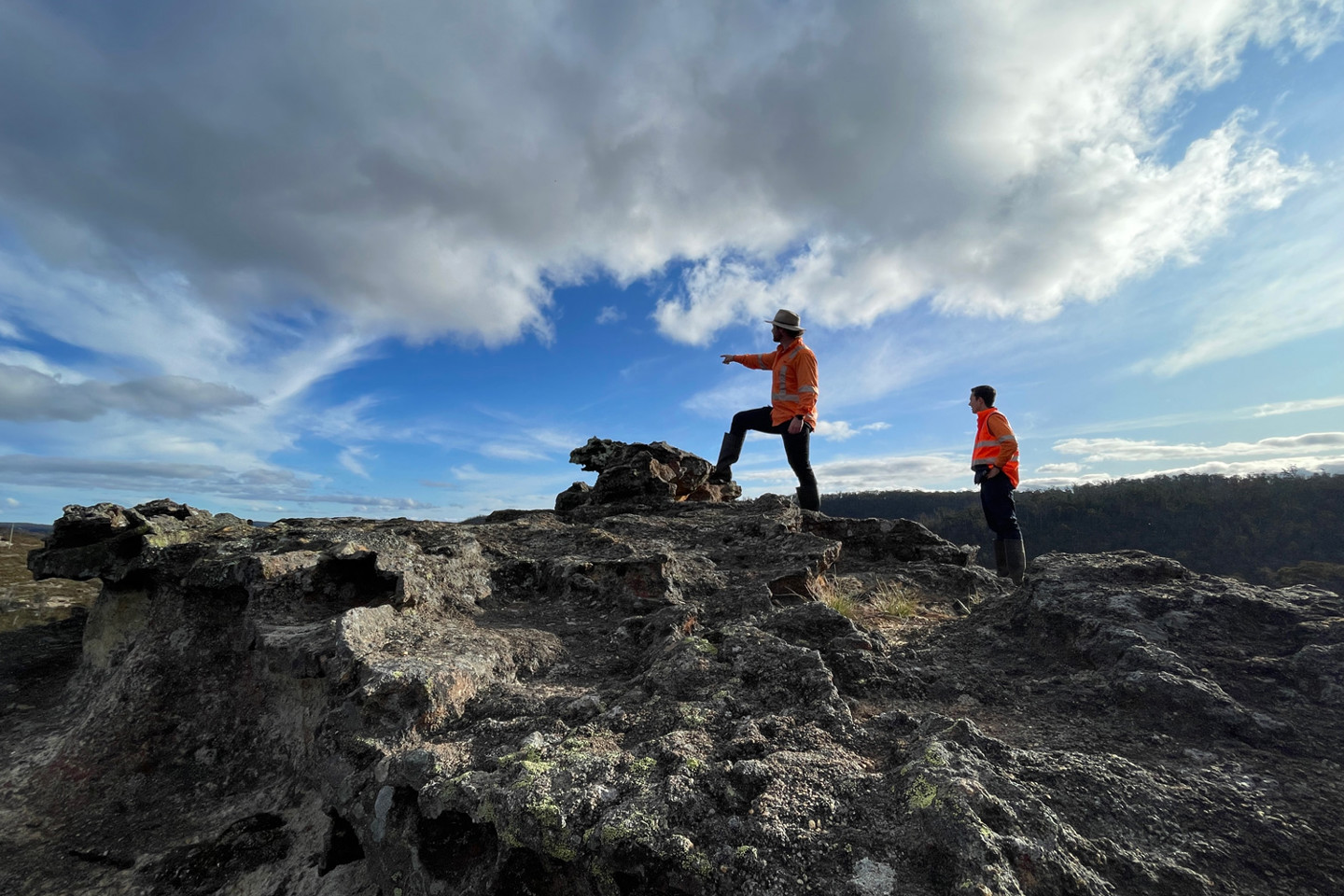 Two RPS environmental scientists dressed in orange high-vis stand on rocky terrain with one pointing to a distance as the clouds sweep overhead. 