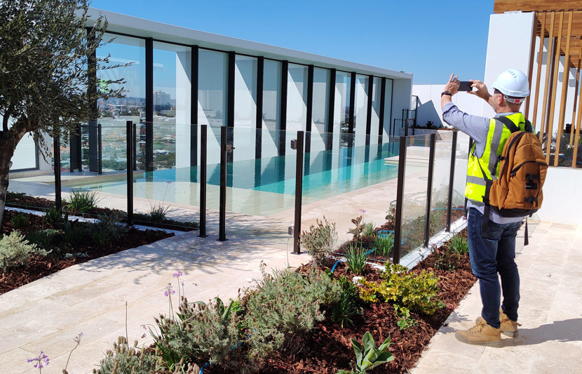 RPS landscape architect dressed in yellow high-vis and hardhat stands in front of an infinity pool and gardens and takes a smart phone photo of the surroundings. 