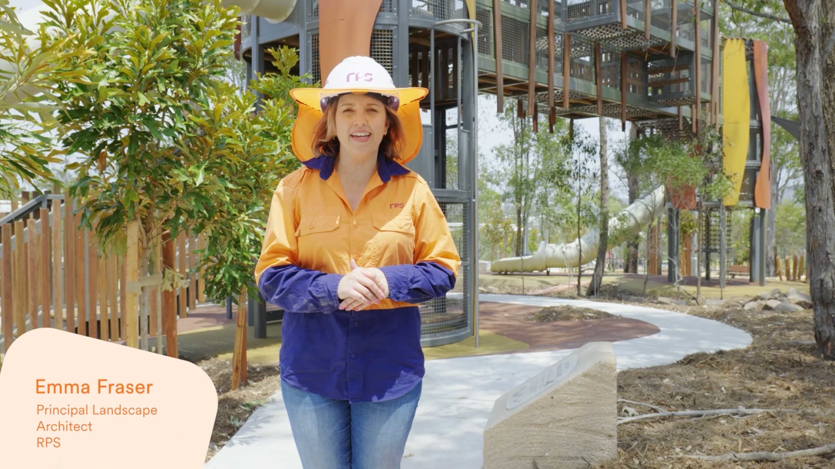 Landscape architect Emma Fraser dons a hardhat featuring the plum RPS logo, and orange and blue PPE gear with jeans underneath and stands in front of a playground with a tube slide.