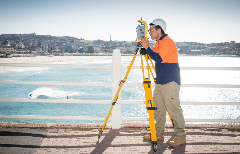 RPS surveyor looks into a surveying tripod with Sydney's Bondi Beach as the backdrop 