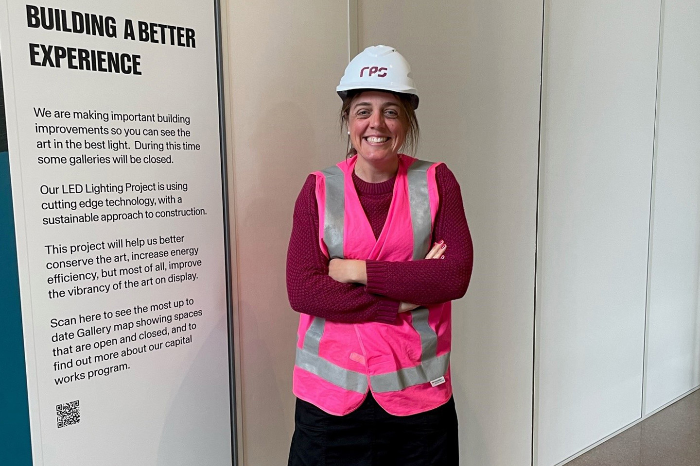 RPS Principal Lead Rebecca Veness wears a logo hard hat with hot pink high-vis and stands in front of a sign at the National Gallery of Australia that reads, 'Building a Better Experience.' 