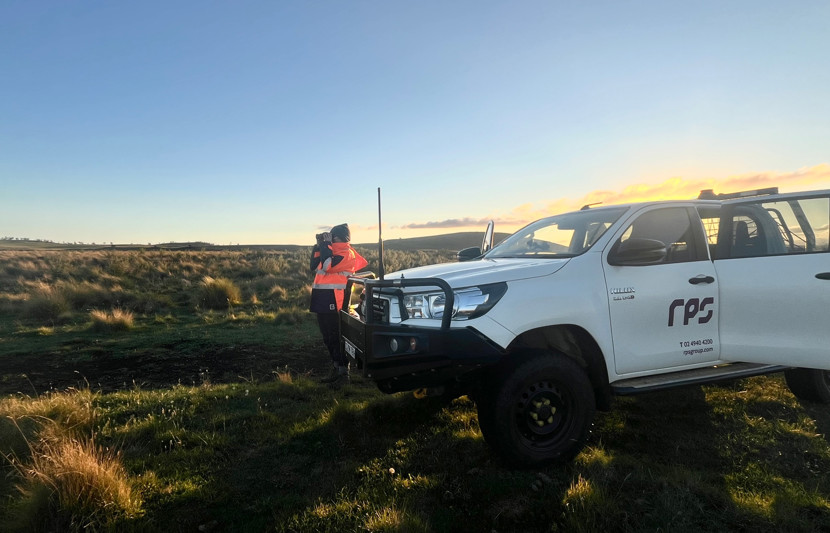 RPS field expert wears orange high-vis PPE and peers through binoculars while surrounded by bushland and leaning on a white RPS branded vehicle during sun-set. 