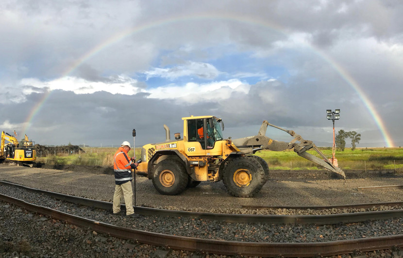 RPS surveyor stands in the middle of railway tracks with a digger in front of him and a large rainbow in the distance. 