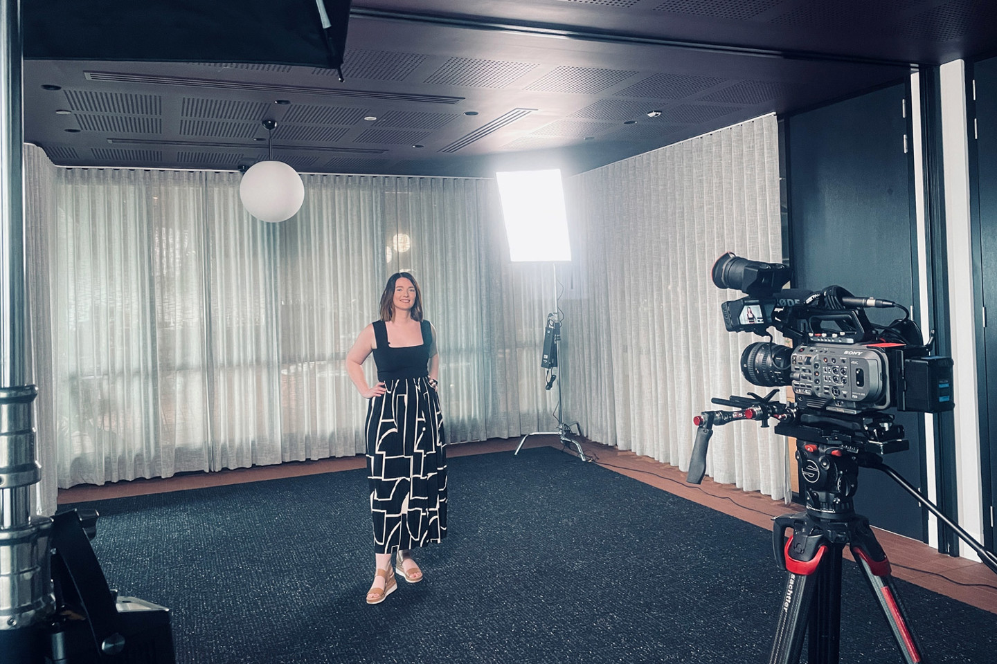 RPS Principal Urban Designer Mel Drake wears a black and white outfit and brown sandals, and is facing a video camera hands on hips and smiling. 