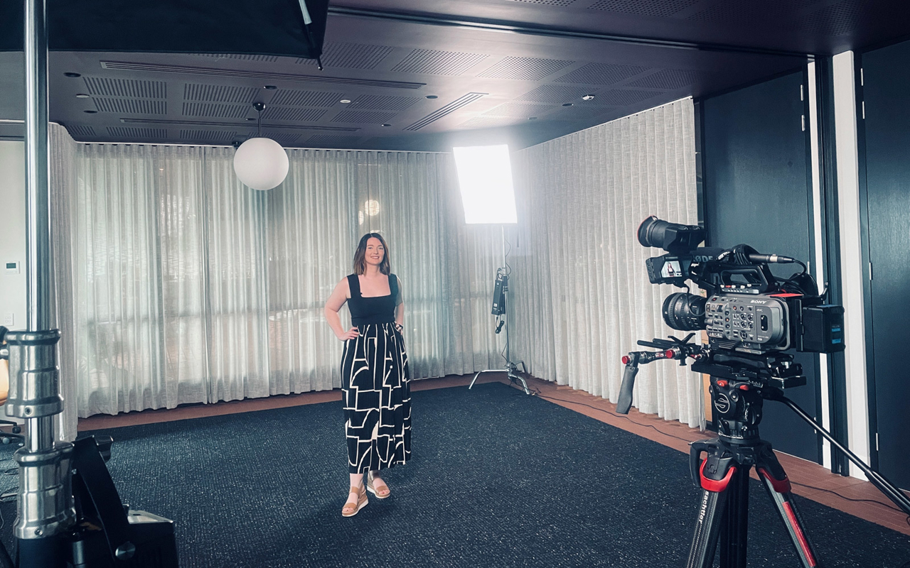 RPS Principal Urban Designer Mel Drake wears a black and white outfit and brown sandals, and is facing a video camera hands on hips and smiling.