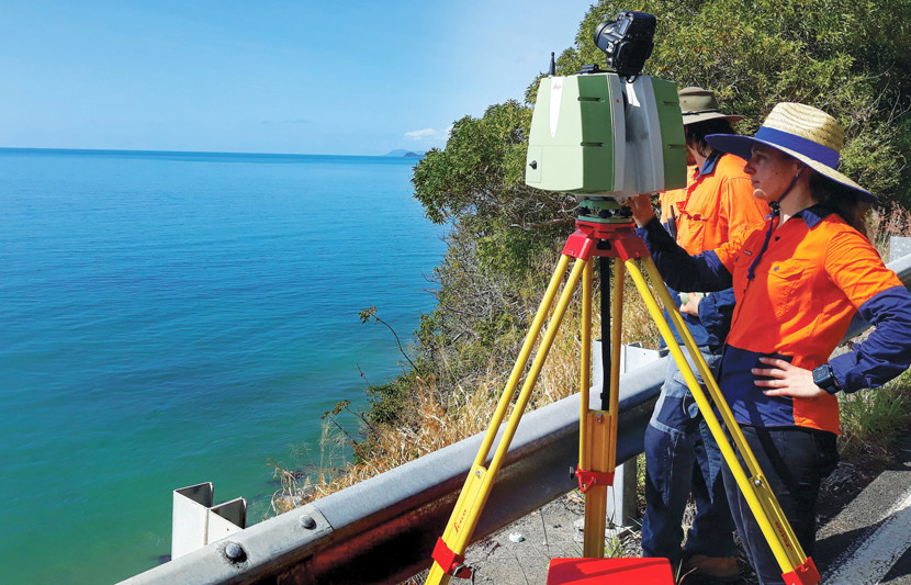 An RPS female surveyor dressed in orange high-vis stands behind surveying equipment with a scrubby bushland to her right and the ocean in front of her with only a road barrier between the water and herself. 